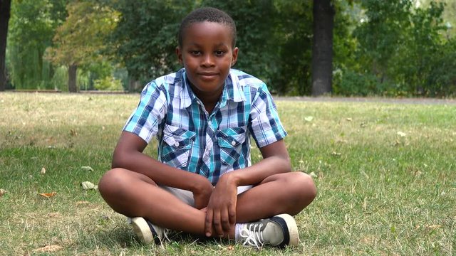 A young  black boy sits on grass in a park and smiles at the camera