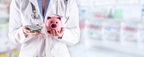 Zelfklevend Fotobehang Doctor woman hands holding pills and piggy bank. Blurred pharmacy interior in the background © weyo