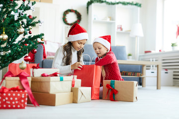 Obraz na płótnie Canvas Siblings in Santa caps looking in red paper bag among giftboxes on the floor