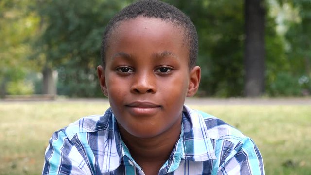 A young  black boy sits on grass in a park and smiles at the camera - face closeup
