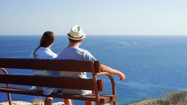 Young couple in love sitting relaxing on bench at edge of cliff. Husband in hat and wife on honeymoon enjoying breathtaking view of blue Mediterranean sea. Concept of freedom and love