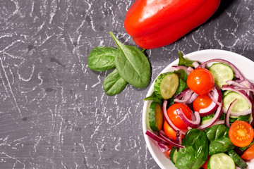 Vegetable salad in a white plate on a gray background, next to the Bulgarian pepper, tomatoes, spinach. Selective focus, space for text.