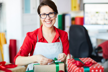 Cheerful young woman with toothy smile looking at you while tying ribbon on giftbox