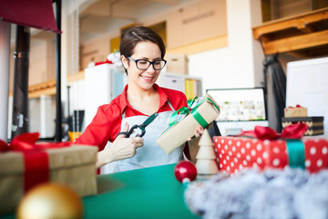 Pretty young designer in workwear sitting in studio and preparing Christmas gifts by workplace