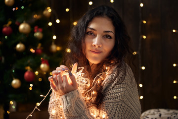 young girl is in christmas lights and decoration, dressed in white, fir tree on dark wooden background, winter holiday concept