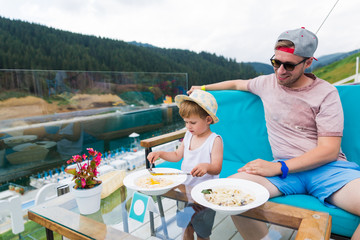 The cute little boy sitting by the table outdoor and eating the pasta and his father sitting near