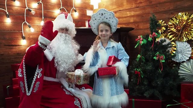 Ded Mroz Santa Claus and the little girl Snegurochka hold boxes with gifts. New Year's Christmas fairy tale characters in the house at a fireplace with smiles welcome the viewer.