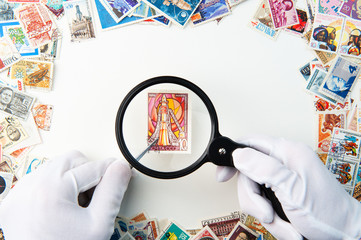 Hands in white gloves holding postage stamp with a forceps and magnifier above the table