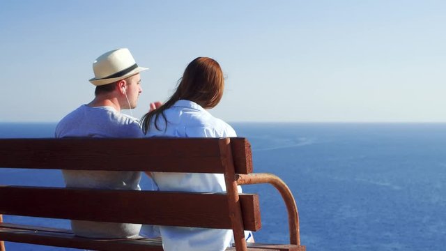 Young couple in love relaxing on bench at edge of cliff listening music with earphones via app on mobile phone. Husband and wife on honeymoon enjoying breathtaking view of blue Mediterranean sea