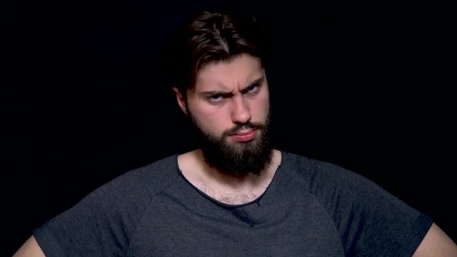 Angry young man with beard makes disagree sign by shaking his head, isolated on black background. Handsome man wearing grey sweater over isolated background looking unhappy and angry showing rejection