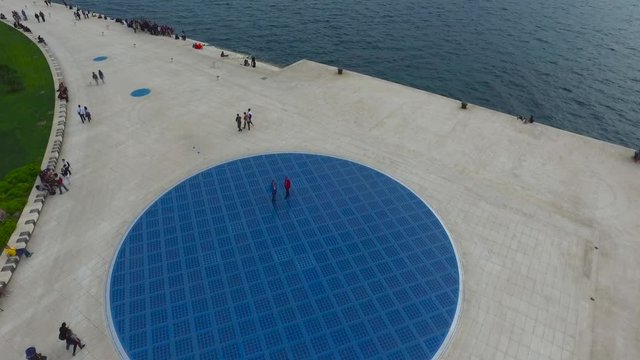 Aerial view of waterfront plaza in Zadar, Croatia with geometric blue circle design and people walking, urban drone footage.