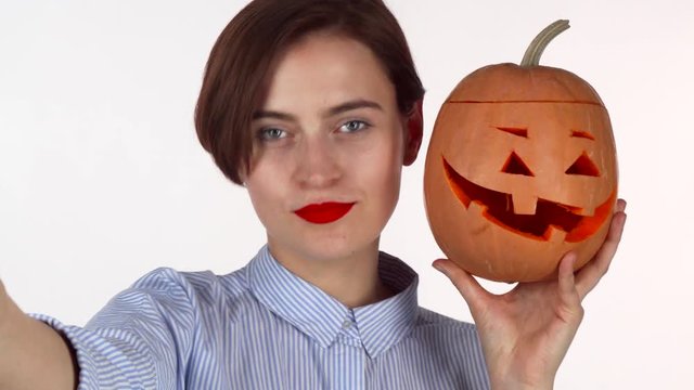 Lovely red lipped woman taking selfies with carved Halloween pumpkin. Cheerful young female enjoying celebrating Halloween, having fun, taking photos of herself. Spooky concept