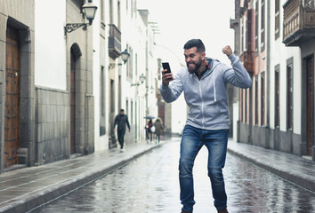 Excited young man looking at cellphone with victorious expression in the middle of the street. Happy hipster with grey hoodie holding mobile device and fist up. Online bet lucky winner concept