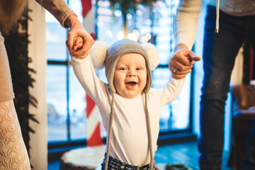 Funny little baby boy 1 year old learning walk home in winter in a decorated New Year house. Young family dad and mom hold by the hands of his son in the loft interior wooden floor near the window