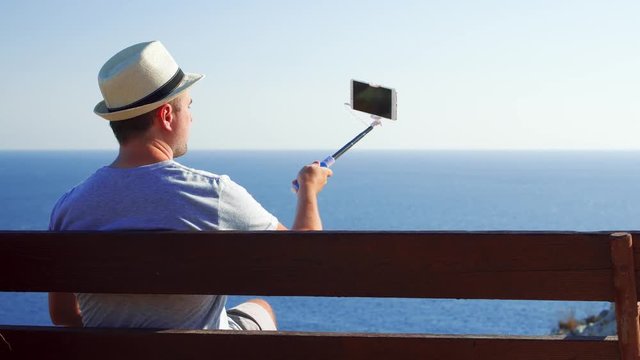 Young handsome man sitting relaxing on bench at edge of cliff doing selfie on cellphone with selfie stick. Male enjoying breathtaking view of blue Mediterranean sea making photos on mobile phone