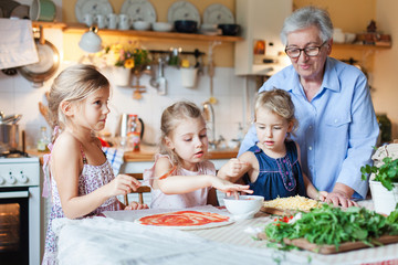 Family and kids cooking pizza in cozy home kitchen. Grandmother and three sisters preparing homemade italian food. Funny little girls helping senior woman, eating ingredients. Children chef concept