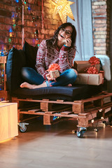 Obraz na płótnie Canvas Cheerful beautiful girl talking by phone with friends while sitting on a couch with gift boxes in a decorated room with loft interior.