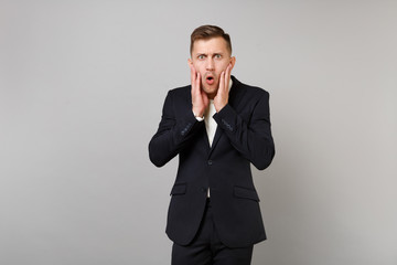 Portrait of shocked young business man in classic black suit, shirt putting hands on cheeks isolated on grey wall background in studio. Achievement career wealth business concept. Mock up copy space.