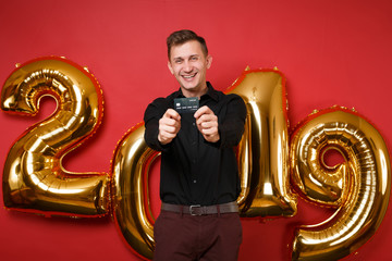 Merry young man in black shirt celebrating holiday party, holding credit card isolated on bright red wall background golden numbers air balloons studio portrait. Happy New Year 2019 Christmas concept.