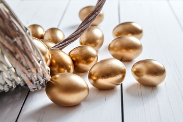 Easter golden eggs spilled out of wicker basket on white wooden table background