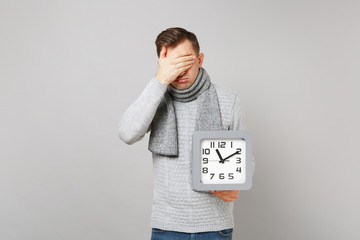 Tired young man in gray sweater, scarf covering face with hand hold square clock isolated on grey...