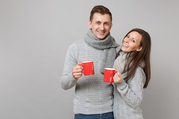 Couple girl guy in gray sweaters, scarves hold cups with tea isolated on grey wall background, studio portrait. Healthy lifestyle, ill sick disease treatment, cold season concept. Mock up copy space.