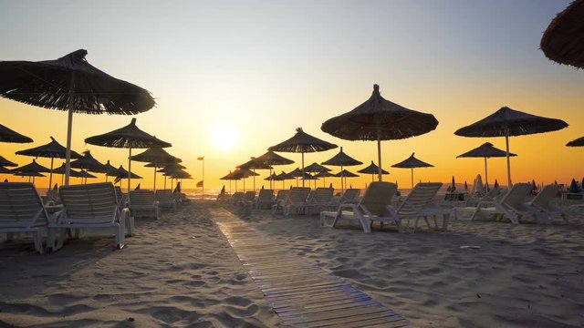 Happy woman in white dress and hat walking and turning on beach at sunset. Seaside resort with sun loungers and wooden sheds against sunset, cinematic
