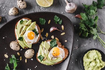 Whole grain bread sandwiches with fried quail egg, avocado, herbs and seeds in plate on grey background. Clean eating, healthy vegan breakfast. Top view, flat lay