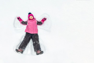 Cute little kid girl in warm ski sport suit making snow angel outdoors. Kid having fun lying on...