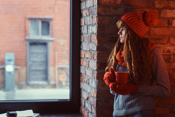 Redhead girl wearing a warm sweater and hat holding a cup of coffee while leaning on a brick wall...