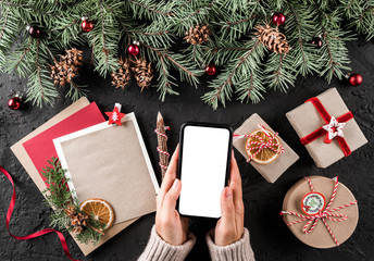 Female hands holding mobile phone. Christmas gift boxes with red decoration, Fir branches, pine cones on holiday background. Xmas and Happy New Year card. Flat lay, top view