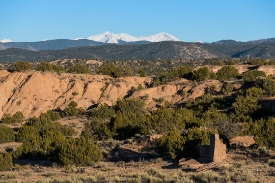 High desert red rock landscape with juniper trees and pinon pines contrasted with the snow-capped peaks of the Sangre de Cristo mountain range in the distance near Santa Fe, New Mexico