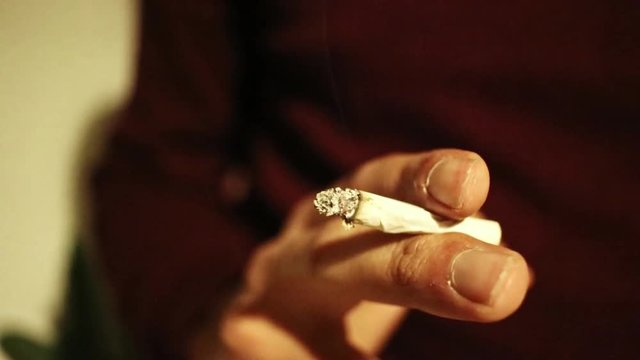 Nahaufnahme von m&auml;nnlicher Hand die selbst gedrehte brennende Zigarette mit aufsteigendem Rauch h&auml;lt