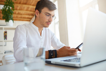 Indoor shot of pensive handsome young businessman wears white shirt and glasses using laptop and mobile phone sitting and working at the table in office