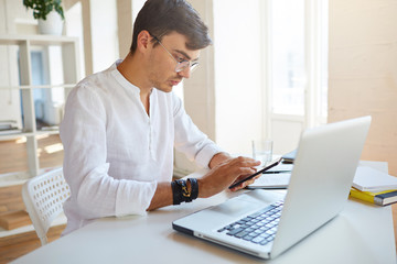 Portrait of concentrated handsome young businessman wears white shirt and spectacles using laptop and smartphone working at the table in office