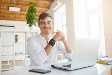 Portrait of cheerful confident young businessman wears white shirt and glasses using laptop and smartphone sitting at the table in office