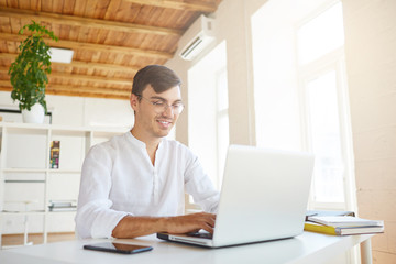 Indoor shot of happy attractive young businessman wears white shirt and glasses feels satisfied, using laptop and smartphone working at the table in office