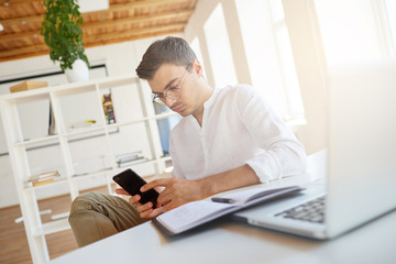 Fototapeta premium Portrait of pensive serious young businessman wears white shirt and glasses working with laptop and using mobile phone at the table in office
