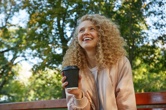 A young beautiful girl blonde sits on a park bench, drinking coffee, looks on a companion, speaking with someone, laughing, feels happy, in a good company, has a good time