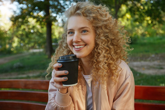 Outdoor photo of young beautiful girl blonde sits on a park bench, drinking coffee, looks on a companion,away, speaking with someone, smiling, feels happy, in a good company, has a good time, dreaming