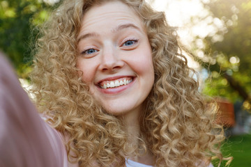 Young beautiful funny cool girl blonde with curly hair, widely smiling and looking into the camera, making selfie, nature background