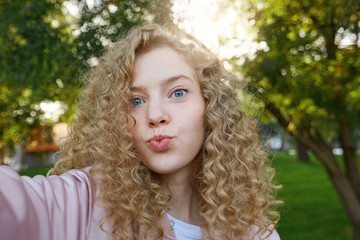 Naklejka premium Beautiful attractive girl blonde with curly hair and blue charming eyes, sending blowing kiss with pout lips looking at camera, making selfie, nature background