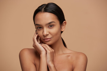 Head shot of an attractive mixed race girl, looking gently and tenderly, demonstrates healthy beautiful skin, calm and pleased, keeps hands near the face, over beige background