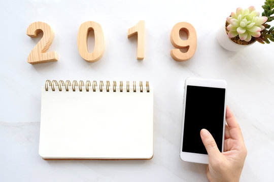 2019 wood letters, hand holding smart phone  with blank screen and blank notebook paper on white marble table background, 2019 new year mock up, template with copy space for text, top view