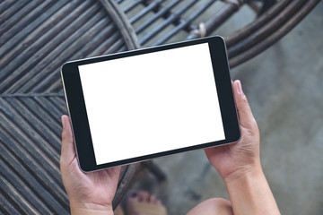 Top view mockup image of hands holding and using black tablet pc with blank white desktop screen while sitting in outdoor