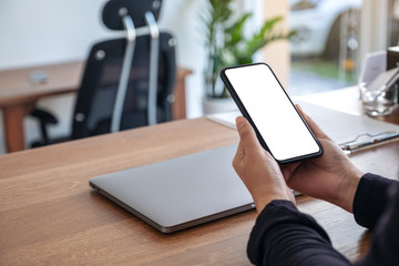 Mockup image of hands holding black mobile phone with blank white screen with laptop on wooden table