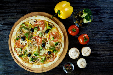 Hot vegetarian pizza and fresh peppers, tomatoes, mushrooms, olives, broccoli and spices close-up on a dark wooden surface.