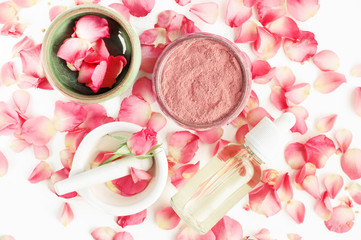 Top view botanical skincare home spa treatment with pink petals, rose blossom, clay face mask, bottle of essential oil.