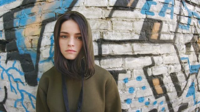 Serious brooding girl in hood standing near wall with graffiti in sports clothes. Portrait of good-looking teen in stylish clothes. Lifestyle. Outdoors. Hip-hop concept.