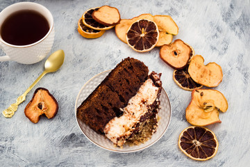 A portion of chocolate cake on a plate. A layer of white meringue, brown dough. Dried slices of lemon, apples, pears. Tea in white mug. Light background.